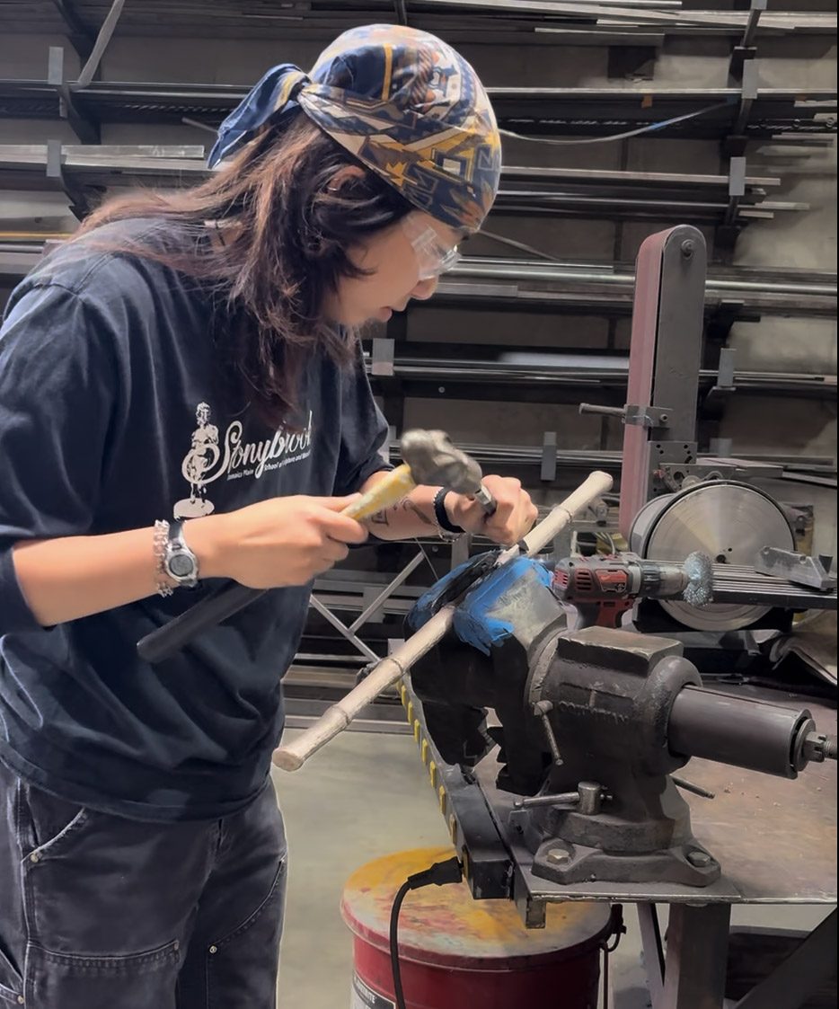 An apprentice hand-finishing a metal rod at a vise in the One Off Furniture workshop