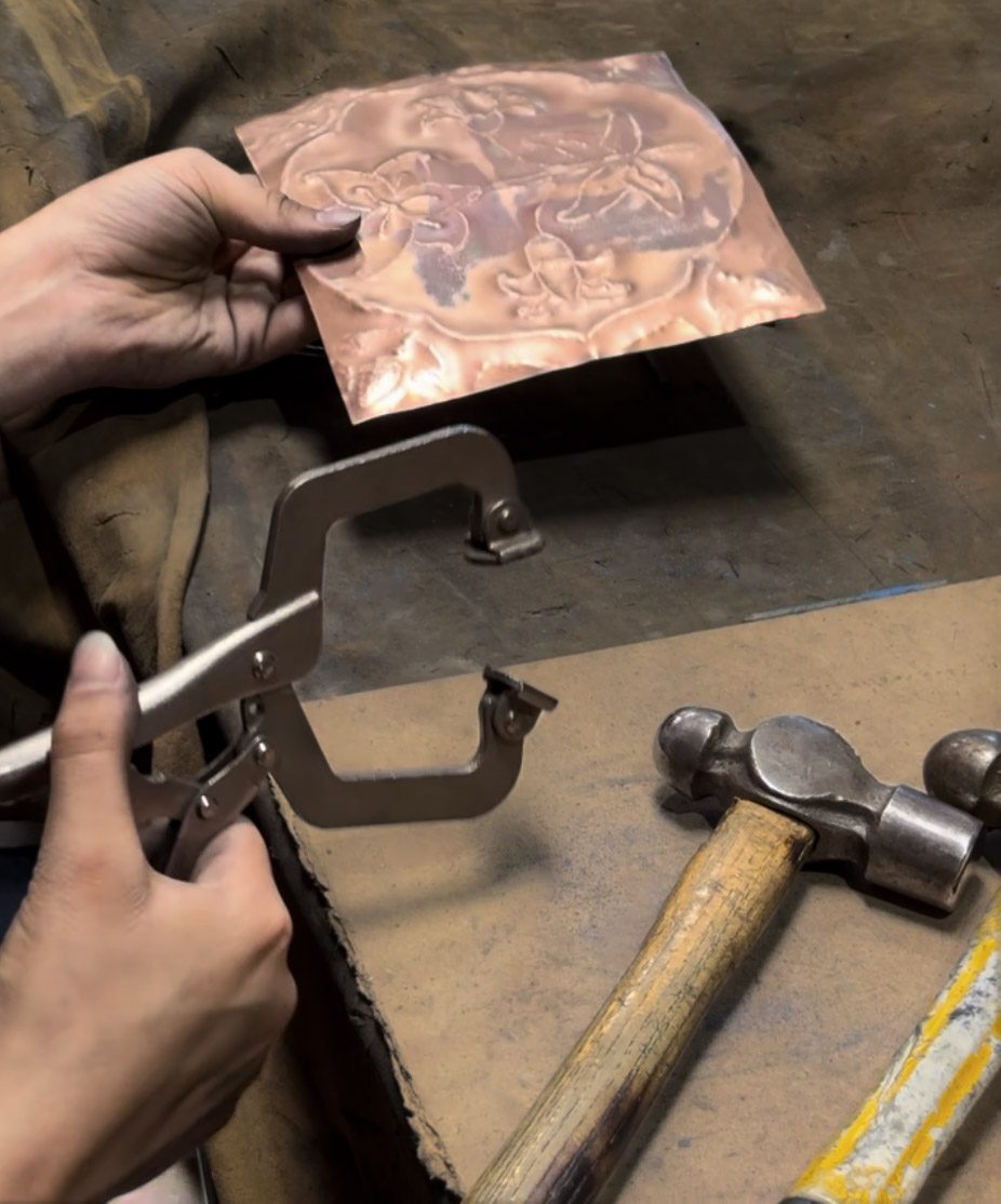Close-up of a hand-tooled copper repoussé panel with a floral design, held above hammers and a bench vise